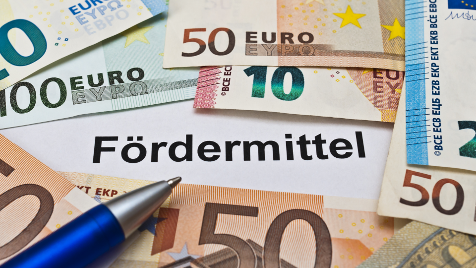 Abgebildet sind mehrere Euro-Geldscheine und ein Kugelschreiber. In der Mitte des Bildes steht "Fördermittel". 