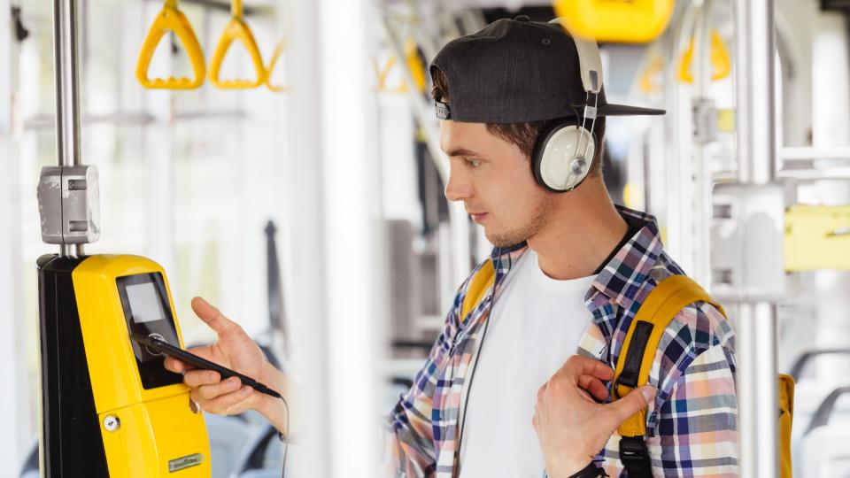 Junger Mann bezahlt mit seinem Handy an einem Ticketautomaten in einer Straßenbahn.