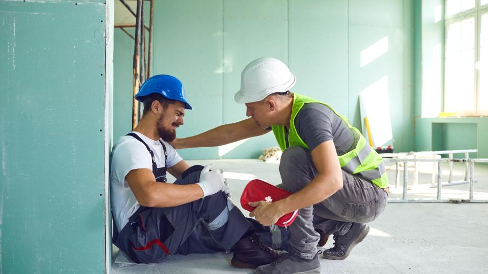 Ein Handwerker auf einer Baustelle hält sich schmerzend das Knie ein Weiterer Handwerker kniet mit einer Erste-Hilfe Tasche vor Ihm
