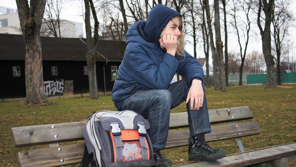 Ein Schüler sitzt alleine und nachdenklich auf einer Parkbank im Freien. Seinen Schulranzen hat er neben sich abgelegt. 